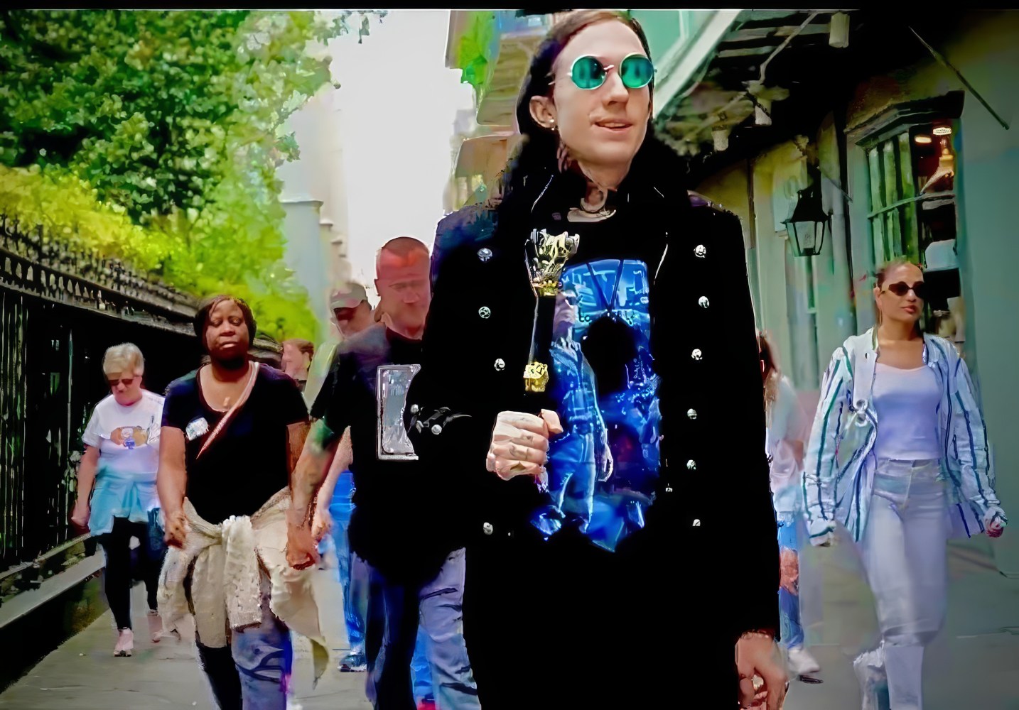 Person in sunglasses and black jacket holding a staff, walking on a city street with others.