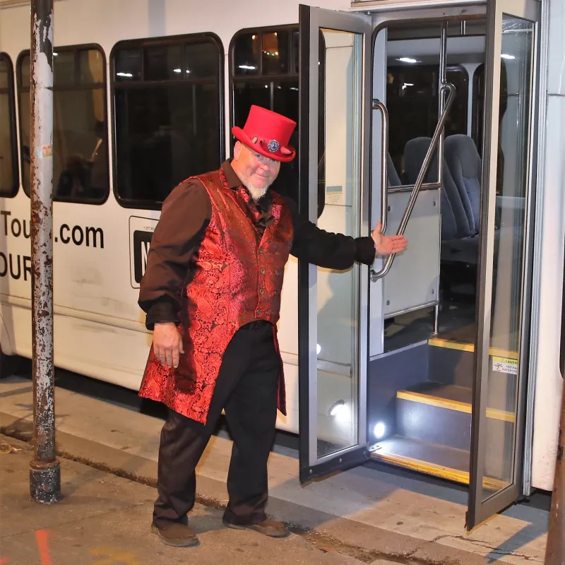 Person in red top hat and vest gesturing at bus entrance in a nighttime setting.